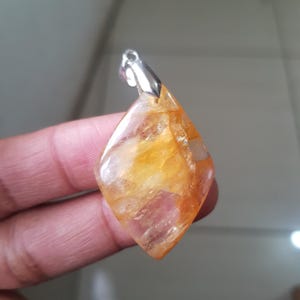 Può includere: Un ciondolo a forma di diamante con una cauzione d'argento. Il ciondolo è una pietra preziosa traslucida e lucidata con sfumature di arancione e giallo. La pietra preziosa è tenuta da una cauzione d'argento.