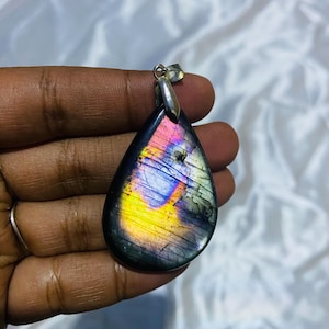 May include: A teardrop-shaped labradorite pendant featuring a polished surface with iridescent flashes of yellow, blue, and purple. The gemstone is set in a simple silver-toned metal bail at the top, showcasing natural internal fractures.