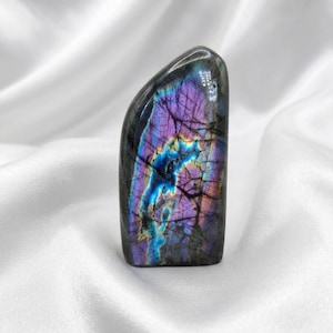 Peut inclure: Cristal de labradorite poli, avec une base sombre et des reflets irisés de violet, bleu et vert. La pierre a un sommet lisse et arrondi et une base rectangulaire. Posé sur un tissu blanc et soyeux.
