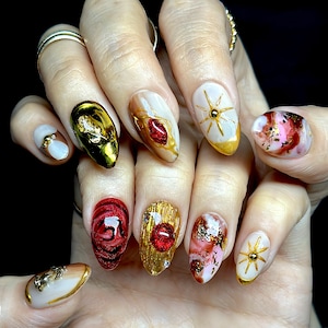 May include: A hand displaying a set of elaborately designed, long, oval-shaped fingernails. The nails feature a variety of designs, including gold accents, red gems, and intricate patterns in shades of gold, red, white, and pink. Some nails have starburst designs.