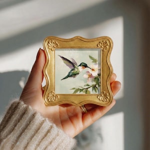 May include: A small square painting of a hummingbird hovering near light pink flowers, encased in an ornate gold-coloured frame with scalloped edges and decorative floral motifs at each corner, featuring visible brushstrokes and soft colours.