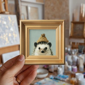May include: A small square painting of a hedgehog wearing a tan knit beanie, set within a decorative gold-toned frame. The hedgehog has white and brown quills, dark eyes, and a black nose, depicted against a pale blue-green background.