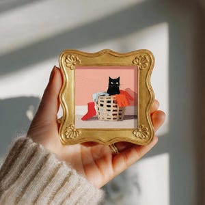 Puede incluir: Un pequeño marco cuadrado dorado y ornamentado que contiene una pintura de un gato negro sentado dentro de una cesta de ropa tejida llena de prendas, incluyendo un calcetín rojo brillante, sobre un fondo rosa claro sólido.
