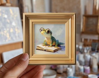 May include: A small square painting in a gold frame depicting a green and yellow lizard wearing round glasses while sitting on an open book. The artwork features soft brushstrokes and a light-colored background, capturing the lizard in a seated pose.