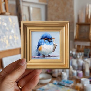 May include: A framed miniature painting of a bluebird wearing orange glasses and holding a book. The bird has white and orange accents and is perched on a branch. The frame is gold.