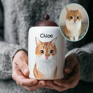 May include: A white ceramic pet urn with a detailed portrait of a tabby cat. The urn has a dark brown wooden lid and the name "Chloe" is written in black script. An oval portrait of the cat is next to the urn.