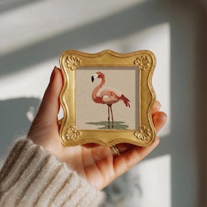 Könnte beinhalten: Ein kleiner quadratischer Bilderrahmen mit metallisch goldener Oberfläche und verzierten gewellten Rändern. Der Rahmen enthält eine gedruckte Illustration eines rosa Flamingos, der in flachem grünem Wasser vor einem strukturierten beigen Hintergrund steht.