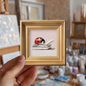 Puede incluir: Pequeña pintura cuadrada enmarcada que representa una mariquita sobre una pila de libros. La mariquita es roja con puntos negros. El marco es dorado. La pintura tiene un estilo suave y pictórico.