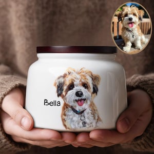 Op de afbeelding: Witte keramische urn voor huisdieren met een donkerbruin deksel. De urn heeft een geschilderd portret van een hond met bruine, witte en zwarte vacht, en de naam "Bella" in zwart schrift. Een ovale inzet toont een foto van de hond.