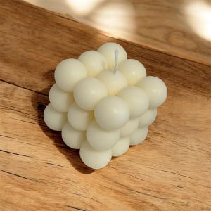 Può includere: Una candela a forma di cubo composta da numerose sfere color avorio. La candela ha una consistenza liscia e un'unica miccia bianca. La candela è posizionata su una superficie di legno.