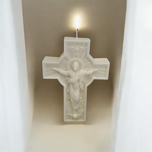 May include: A white, cross-shaped candle featuring a detailed relief of Jesus Christ with outstretched arms and a dove above. The candle is lit, with a visible flame at the top. The background is a neutral beige.