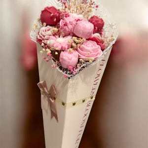 May include: A bouquet of pink and red flowers, including roses and peonies, presented in a white cone-shaped container. The container is decorated with a ribbon and the text "FLOWERS BLOOM FOR".