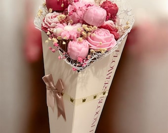 May include: A bouquet of pink and red flowers, including roses and peonies, arranged in a white cone-shaped container. The container is decorated with a ribbon and the text "FLOWERS BLOOM FOR".
