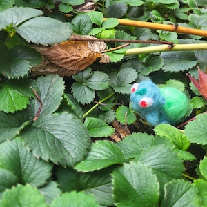 May include: A handmade felt creature with blue and green body, red eyes, and white accents rests among green strawberry plant leaves. The whimsical figure is nestled among the foliage, creating a natural setting.