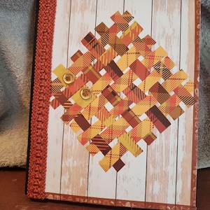 May include: A handmade journal with a decorative woven plaid design in shades of orange, yellow, and brown. The journal features two round, tan buttons and a rust-coloured trim along the spine. The background has a wood grain pattern.