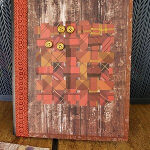 May include: A handcrafted journal with a wood-effect cover. The cover has a plaid pattern in red, orange, and brown, with three gold buttons. The spine is bound with decorative orange trim.