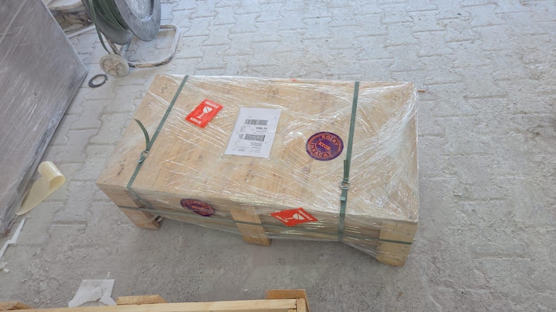 Puede incluir: Una caja de env&iacute;o rectangular de madera asegurada con correas de pl&aacute;stico verde y envuelta en film pl&aacute;stico transparente. La caja presenta una etiqueta de env&iacute;o blanca, dos pegatinas rojas de advertencia de fr&aacute;gil y dos etiquetas circulares.