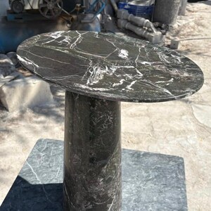 May include: A dark marble side table with a round top and a cylindrical base. The table's surface is a dark gray with white veining. The base is a solid, dark gray color. The table is approximately 24 inches tall.