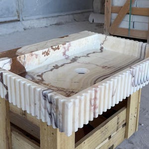 May include: Rectangular onyx bathroom sink with a light beige and brown marbled pattern. The sink has a ridged, vertical design around the edges. It is set on a wooden pallet.