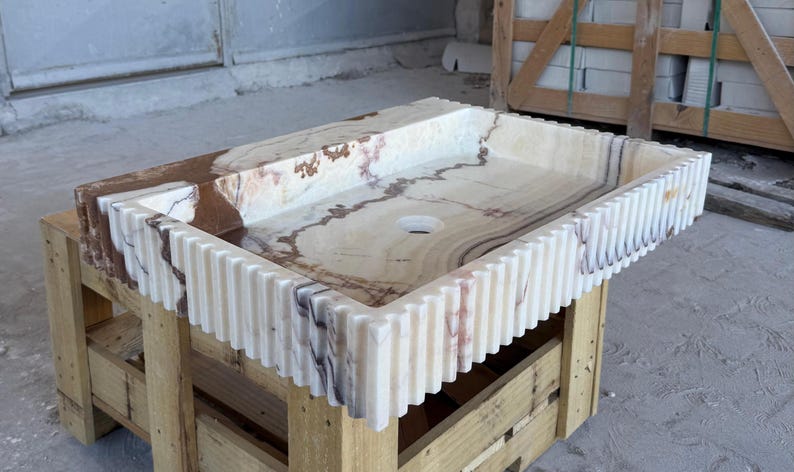 May include: Rectangular onyx bathroom sink with a light beige and brown marbled pattern. The sink has a ridged, vertical design around the edges. It is set on a wooden pallet.