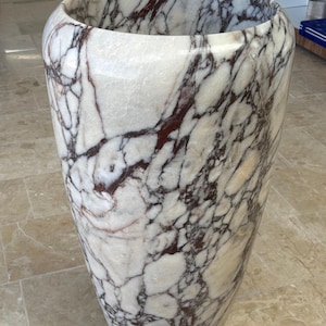 May include: A tall, decorative vase with a wide opening, crafted from white marble with striking burgundy veining. The vase has a smooth, polished finish and a rounded shape, suitable for home decor.