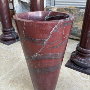 May include: A tall, red marble vase with a flared top and a tapered body. The vase features dark gray and white veining throughout the polished surface. The vase is a decorative item, possibly for floral arrangements.