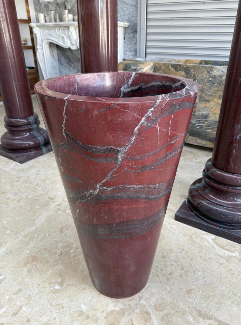 May include: A tall, red marble vase with a flared top and a tapered body. The vase features dark gray and white veining throughout the polished surface. The vase is a decorative item, possibly for floral arrangements.