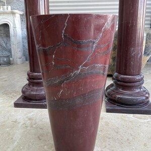 May include: A tall, red marble vase with a flared top and a dark, veined pattern. The vase is set in front of two matching red marble columns. The columns have a square base and a round top.