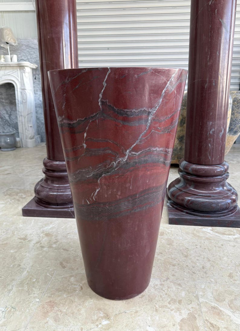 May include: A tall, red marble vase with a flared top and a dark, veined pattern. The vase is set in front of two matching red marble columns. The columns have a square base and a round top.