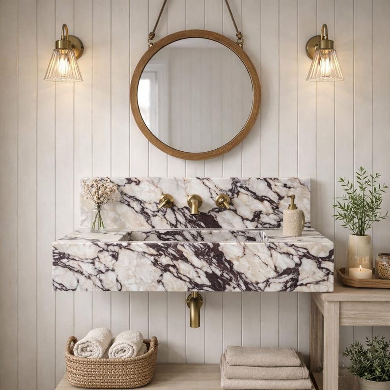 Puede incluir: Un lavabo rectangular de m&aacute;rmol montado en la pared con vetas de color p&uacute;rpura intenso y blanco, que cuenta con un grifo de lat&oacute;n y accesorios a juego. Sobre el lavabo cuelga un espejo redondo con marco de madera, flanqueado por dos apliques de lat&oacute;n.