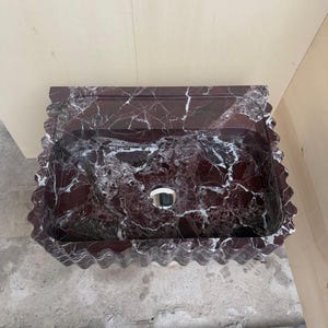 May include: A rectangular, burgundy marble sink with white veining. The sink has a wavy, scalloped edge and a circular drain. The sink is made of a polished stone material, and is designed for bathroom use.