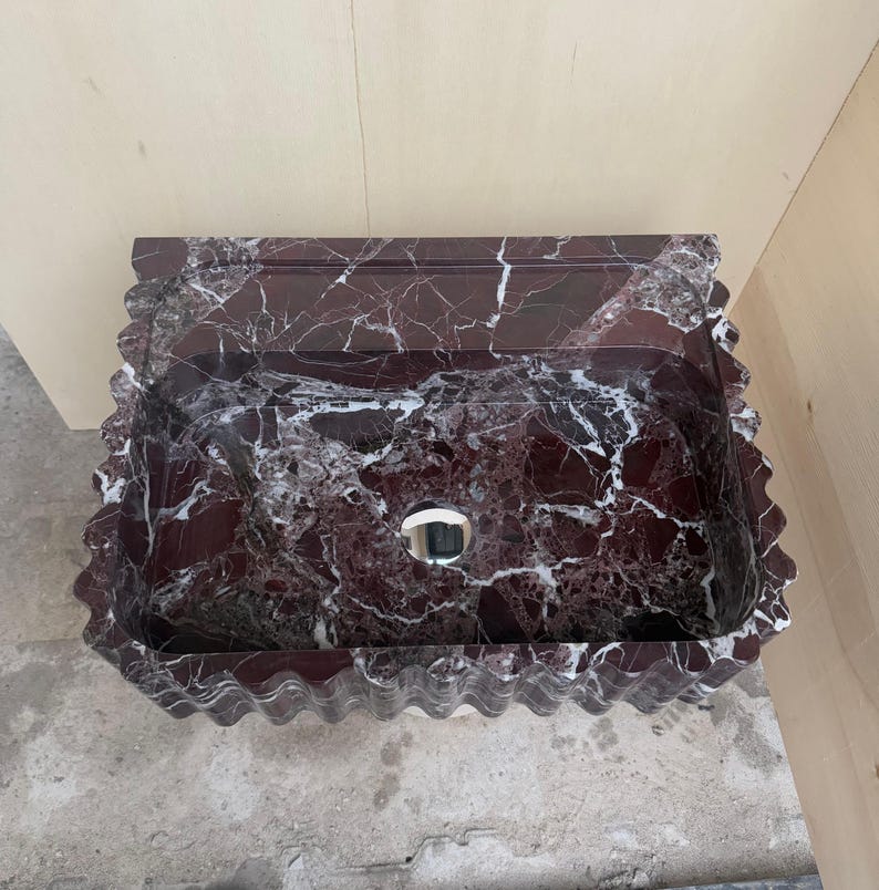 May include: A rectangular, burgundy marble sink with white veining. The sink has a wavy, scalloped edge and a circular drain. The sink is made of a polished stone material, and is designed for bathroom use.