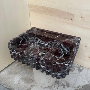 May include: A rectangular, burgundy marble sink with white veining. The sink has a wavy, textured edge and a circular drain hole. It is mounted on a cylindrical, light-colored pedestal. The background is a light wood panel.