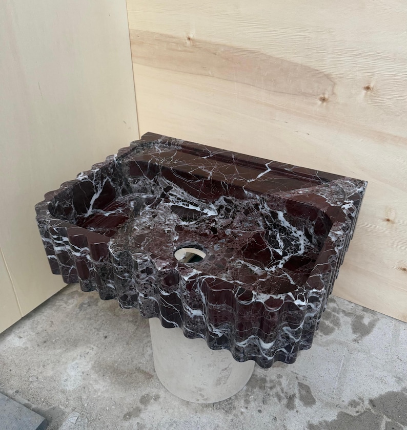 May include: A rectangular, burgundy marble sink with white veining. The sink has a wavy, textured edge and a circular drain hole. It is mounted on a cylindrical, light-colored pedestal. The background is a light wood panel.