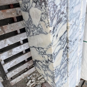 Puede incluir: Un bloque rectangular de m&aacute;rmol blanco pulido que presenta vetas irregulares de color gris oscuro y negro en toda su superficie. La losa de piedra muestra un acabado de alto brillo con un patr&oacute;n natural complejo de dep&oacute;sitos minerales.