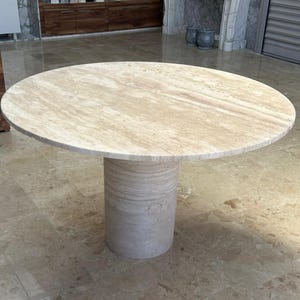 Pode incluir: Mesa redonda de pedra bege com base cilíndrica. O tampo da mesa tem aproximadamente 122 cm de diâmetro. A mesa tem uma superfície natural e texturizada e é adequada para uso interno.