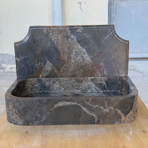 May include: A rectangular, dark gray marble sink with a matching backsplash. The sink has rounded corners and a natural stone pattern with shades of gray, brown, and white. The backsplash has a decorative, curved top edge.