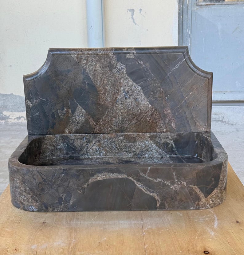 May include: A rectangular, dark gray marble sink with a matching backsplash. The sink has rounded corners and a natural stone pattern with shades of gray, brown, and white. The backsplash has a decorative, curved top edge.
