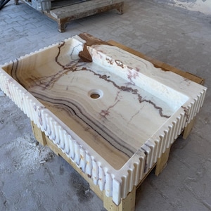 May include: Rectangular onyx bathroom sink with a light beige and brown marbled pattern. The sink has a built-in ledge and a decorative, ridged edge. The sink is set on a wooden support.