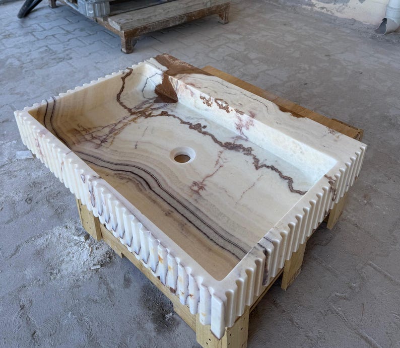 May include: Rectangular onyx bathroom sink with a light beige and brown marbled pattern. The sink has a built-in ledge and a decorative, ridged edge. The sink is set on a wooden support.
