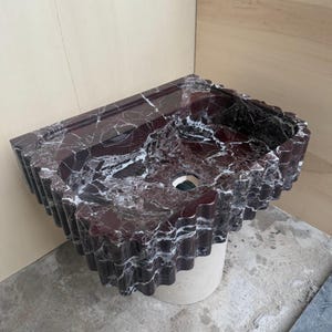 May include: A burgundy marble sink with white veining. The sink has a unique, wavy edge design and a circular drain opening. It is mounted on a light-colored, cylindrical base. The sink's surface is polished, reflecting light.