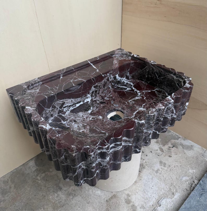 May include: A burgundy marble sink with white veining. The sink has a unique, wavy edge design and a circular drain opening. It is mounted on a light-colored, cylindrical base. The sink's surface is polished, reflecting light.
