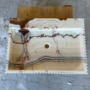 May include: Rectangular onyx bathroom sink with a scalloped edge. The sink features a cream-colored base with brown and black veining. A circular drain hole is centered in the basin. The sink is set against a wooden structure.
