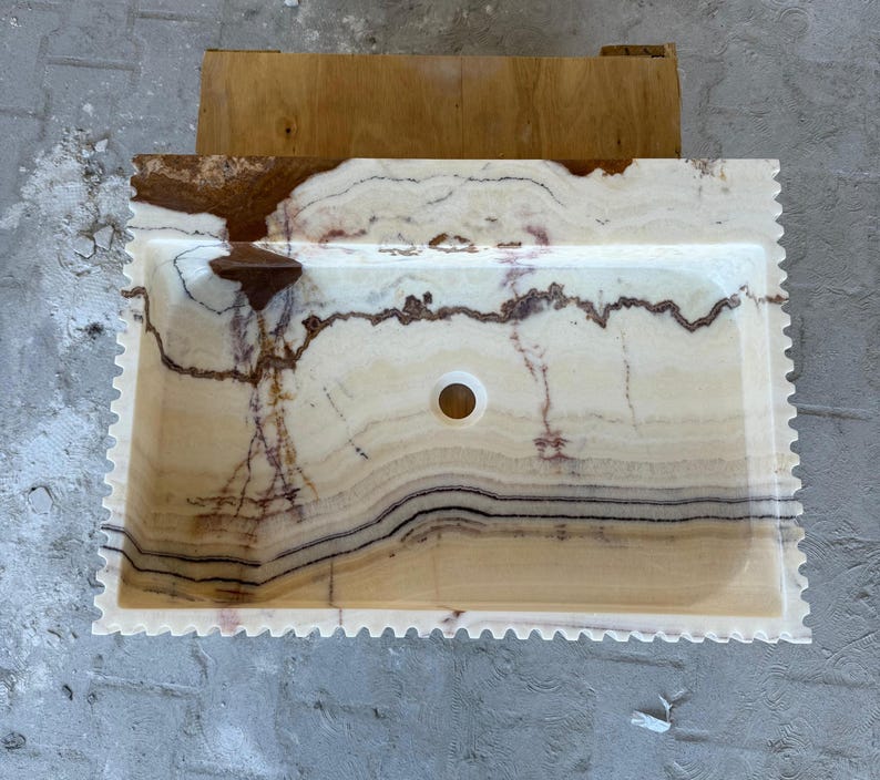 May include: Rectangular onyx bathroom sink with a scalloped edge. The sink features a cream-colored base with brown and black veining. A circular drain hole is centered in the basin. The sink is set against a wooden structure.