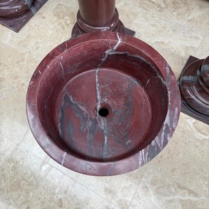 May include: A round, red marble sink with a central drain hole. The sink has a marbled pattern with gray and white veining. The sink is part of a larger marble structure, with a cylindrical column and base visible in the background.