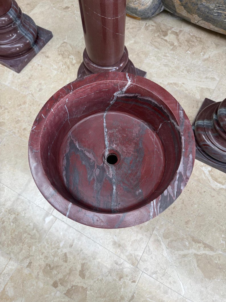May include: A round, red marble sink with a central drain hole. The sink has a marbled pattern with gray and white veining. The sink is part of a larger marble structure, with a cylindrical column and base visible in the background.
