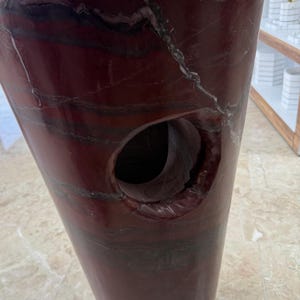 May include: A tall, burgundy-colored stone vase with a large, circular opening. The vase has a polished finish and features natural veining in shades of white and black. The design is a modern, sculptural piece.