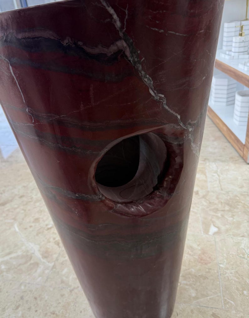 May include: A tall, burgundy-colored stone vase with a large, circular opening. The vase has a polished finish and features natural veining in shades of white and black. The design is a modern, sculptural piece.