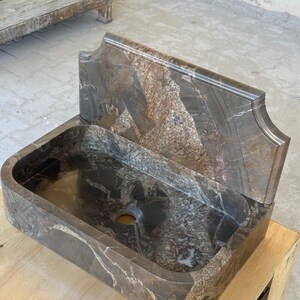 May include: A rectangular, brown marble sink with a matching backsplash. The sink has a rounded basin and a single drain hole. The backsplash has a decorative, curved top edge. The marble has a natural, veined pattern.