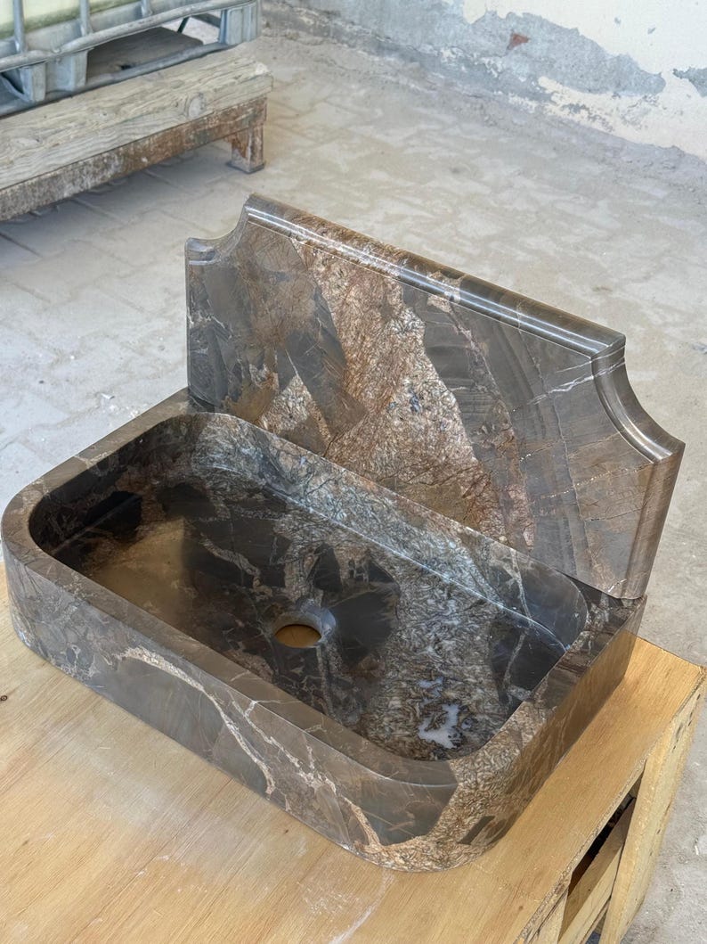 May include: A rectangular, brown marble sink with a matching backsplash. The sink has a rounded basin and a single drain hole. The backsplash has a decorative, curved top edge. The marble has a natural, veined pattern.